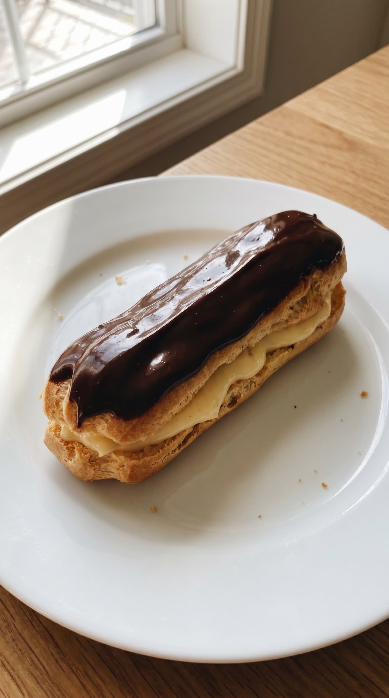 A classic chocolate eclair filled with pastry cream and topped with glossy chocolate glaze on a white plate.