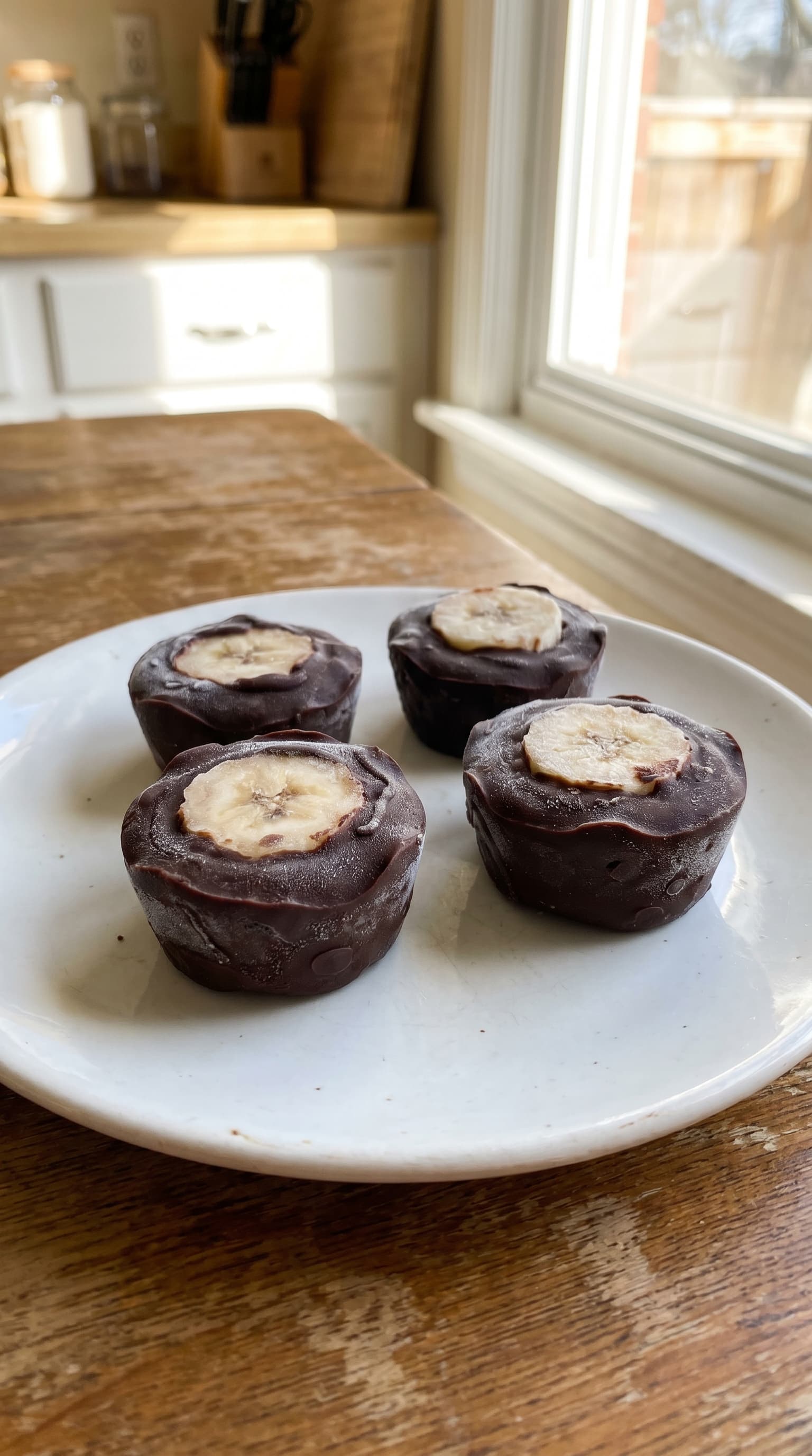 Frozen chocolate banana bites topped with banana slices served on a white plate by a sunny kitchen window.